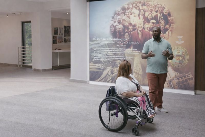 A woman in a wheelchair and a man stand together smiling at the DuSable Museum of African American History in front of a mural depicting the history of Harold Washington