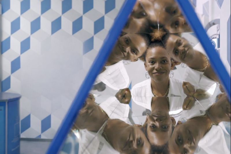 A Deaf woman in a white blazer stands in a kaleidoscope mirror at Chicago's Museum of Illusions, her image reflected multiple times in a circular pattern with blue geometric patterns in the background